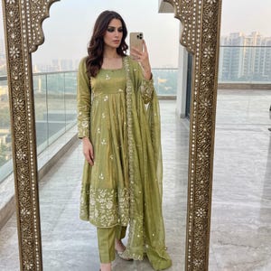 May include: An olive green long-sleeved tunic dress with a square neckline, featuring white floral embroidery along the hem and sleeves, paired with matching straight-leg trousers and a sheer, lightweight olive green dupatta with embroidered edges.
