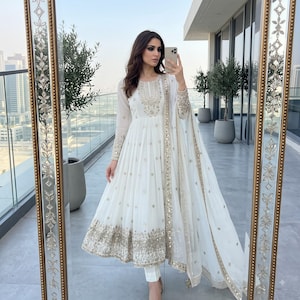 May include: A white floor-length Anarkali dress featuring intricate gold embroidery and mirror work on the bodice, sleeves, and hem. The ensemble includes a matching sheer dupatta with a decorative gold border and slim-fit white trousers.