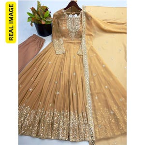 May include: A beige floor-length anarkali dress featuring long sleeves, a round neckline with intricate silver embroidery, and a flared skirt adorned with scattered floral motifs and a wide, heavily embellished silver border at the hem.