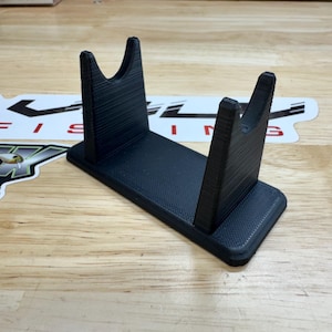 May include: A black, 3D-printed fishing rod holder. The holder has a rectangular base with two upright supports, each with a curved top. The textured surface and the design suggest a functional and durable product.