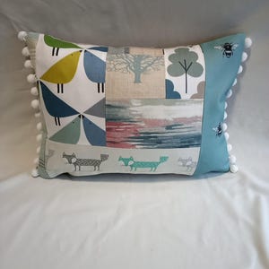 May include: A rectangular cushion with a patchwork design featuring various patterns, including birds, trees, foxes, and bees. The cushion has a light blue background and white pom-poms around the edges.