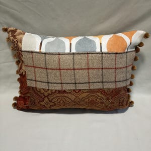 May include: A rectangular cushion with a patchwork design. The cushion has a brown and white checked fabric, a grey and orange geometric fabric, and a brown and gold paisley fabric. The cushion has a fringe border with pom-poms.