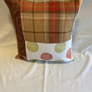 May include: A decorative throw pillow with a patchwork design. The pillow features a brown and red plaid fabric, a gold paisley fabric, and a white fabric with embroidered circles in shades of green and orange.