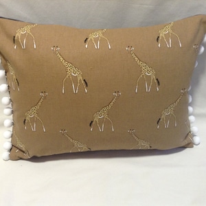 May include: A rectangular cushion in tan fabric, patterned with giraffes. The cushion has white pom-poms along the edges. The giraffes are brown with white and black markings. The cushion is approximately 40 cm x 60 cm.