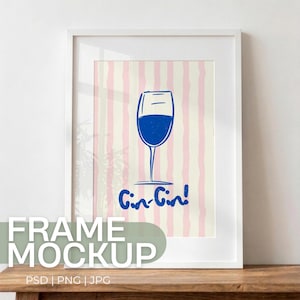 May include: White framed art print featuring a blue wine glass illustration with the words "Gin-Gin!" on a pink and cream striped background. The artwork is displayed on a wooden surface.