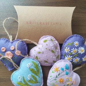 Puede incluir: Un conjunto de adornos de fieltro en forma de corazón en tonos morado, azul y lavanda, cada uno adornado con diseños florales bordados. Los adornos están ensartados en hilo y se presentan con una caja de cartón marrón. El texto "KOPILKABYTANIA" está impreso en la caja.