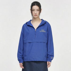 Op de afbeelding: Een vrouw draagt een blauwe anorak met capuchon met de tekst "KEEP SWIMMING" en een donkerblauwe spijkerbroekrok. Ze draagt ook zwart-witte sneakers. De outfit staat tegen een witte achtergrond.