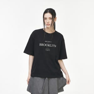 May include: A black t-shirt with the text "MEET ME IN BROOKLYN" in white. Below the text are the coordinates 40.6700° N and 73.9442° W. The shirt has short sleeves and a relaxed fit.