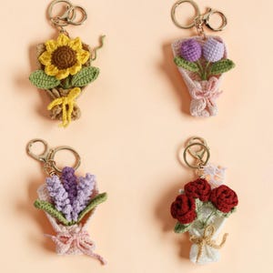 May include: Four crochet flower bouquet keychains. Each keychain features a different flower arrangement: a sunflower, purple tulips, lavender, and red roses. The bouquets are wrapped in paper and tied with a ribbon or twine, and attached to a gold-colored keychain.