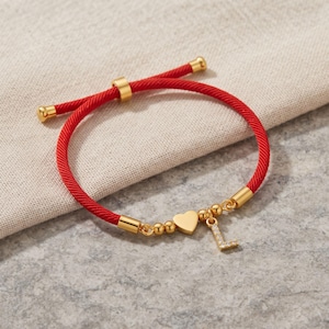 Puede incluir: Una pulsera de cordón rojo con un cierre deslizante dorado, un pequeño dije de corazón dorado y un colgante con la letra L dorada incrustada con pequeñas piedras transparentes. La pulsera tiene terminales metálicos dorados en los extremos.