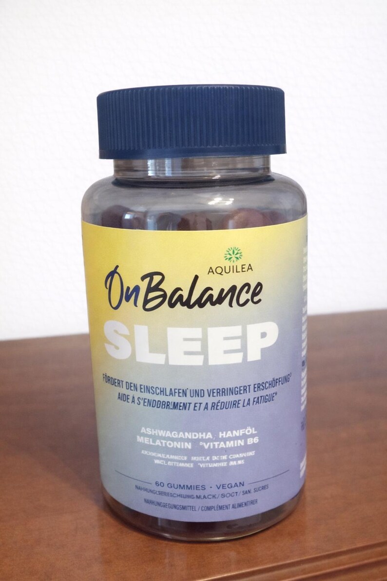 May include: A clear plastic bottle with a dark blue lid, containing gummy vitamins. The label features the words "On Balance SLEEP" in large letters, with additional text in German and French. The bottle is on a wooden surface.