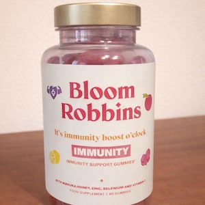 May include: A clear plastic bottle with a gold lid, filled with red gummies. The label reads "Bloom Robbins" and "It's immunity boost o'clock". The label also says "IMMUNITY" and "IMMUNITY SUPPORT GUMMIES".