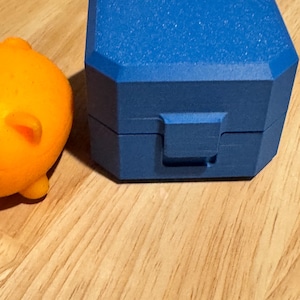 May include: A bright blue, octagonal-shaped storage box with a textured surface and a central latch mechanism, positioned next to a portion of a smooth, rounded orange object. The box features a lid that sits flush with the base.