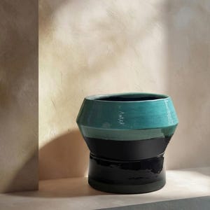 Può includere: Un vaso decorativo in ceramica dal design geometrico. Il vaso presenta una parte superiore turchese, una sezione centrale nera e una base nera. Il vaso è esposto su una superficie di colore neutro.