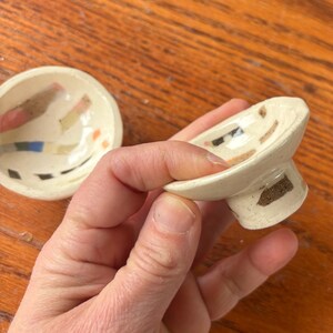 May include: Two small, handmade ceramic bowls. One is a shallow, round bowl with a flat base. The other is a small bowl with a pedestal base. Both bowls are off-white with colorful abstract designs in pink, brown, blue, and orange.