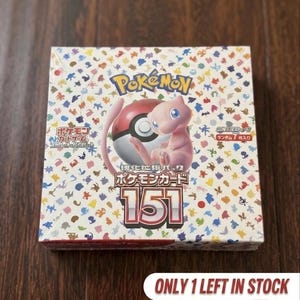 Peut inclure: Une boîte de boosters de cartes à collectionner Pokémon 151 scellée, illustrée avec le personnage Mew rose et une Poké Ball rouge et blanche. La boîte présente un motif coloré de petites silhouettes Pokémon et le texte Pokémon, 151 et des caractères japonais.