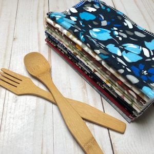 Puede incluir: Una pila de servilletas de tela estampadas, la superior con un dise&ntilde;o floral azul, junto a un tenedor y una cuchara de bamb&uacute;. Las servilletas muestran diversos patrones y colores, incluyendo azul, rojo y blanco. Los cubiertos de madera son de color marr&oacute;n claro.