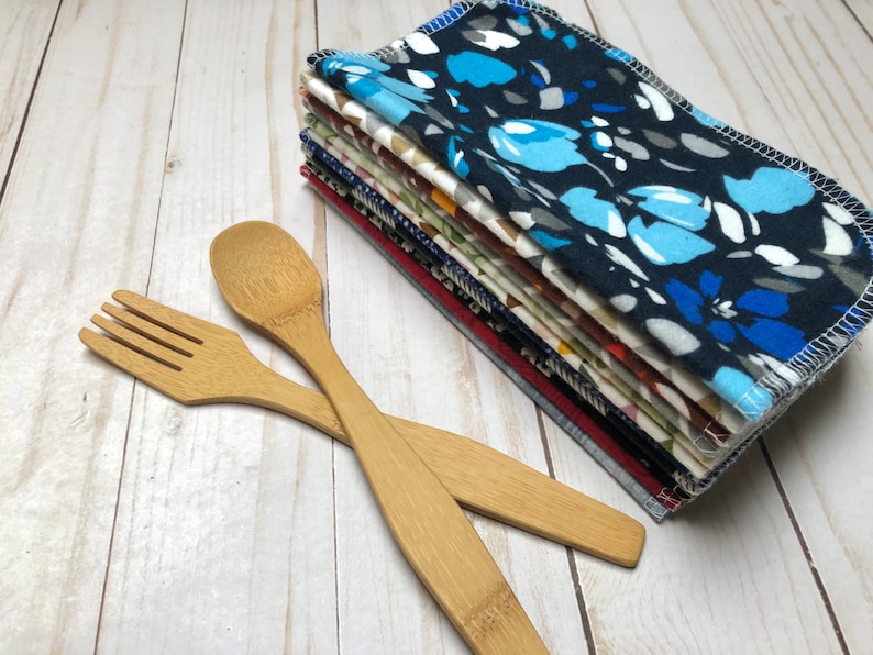 Puede incluir: Una pila de servilletas de tela estampadas, la superior con un dise&ntilde;o floral azul, junto a un tenedor y una cuchara de bamb&uacute;. Las servilletas muestran diversos patrones y colores, incluyendo azul, rojo y blanco. Los cubiertos de madera son de color marr&oacute;n claro.