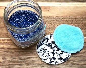 May include: A clear glass jar filled with reusable facial rounds. The rounds feature various patterns, including a blue and white dotted design, a black and white floral print, and a solid light blue terry cloth. The jar sits on a wooden surface.