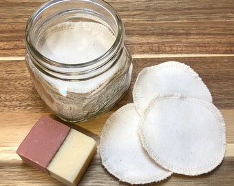 May include: A glass jar filled with reusable cotton rounds, three additional rounds are on a wooden surface next to the jar, and a bar of soap with a brown and white color scheme is also on the surface.