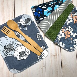 Puede incluir: Dos juegos de servilletas de tela reutilizables. Un juego presenta un estampado floral azul con una cuchara y un tenedor de madera. El otro juego tiene una variedad de patrones que incluyen dise&ntilde;os azules, grises y verdes. Las servilletas est&aacute;n sobre una superficie de madera clara.