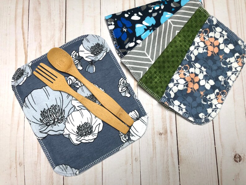 Puede incluir: Dos juegos de servilletas de tela reutilizables. Un juego presenta un estampado floral azul con una cuchara y un tenedor de madera. El otro juego tiene una variedad de patrones que incluyen dise&ntilde;os azules, grises y verdes. Las servilletas est&aacute;n sobre una superficie de madera clara.