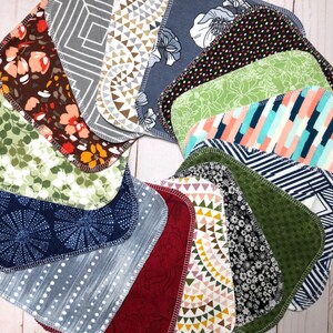 Puede incluir: Una colecci&oacute;n de baberos de tela para beb&eacute;s, dispuestos en un patr&oacute;n circular. Los baberos presentan varios dise&ntilde;os, incluyendo florales, geom&eacute;tricos y abstractos, en colores como verde, azul, marr&oacute;n y blanco. Cada babero tiene una forma redondeada y un borde cosido en contraste.