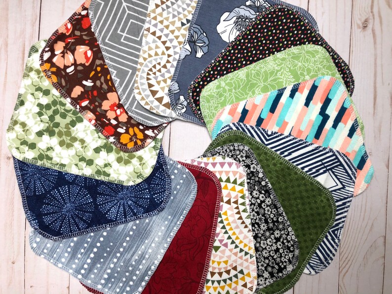 Puede incluir: Una colecci&oacute;n de baberos de tela para beb&eacute;s, dispuestos en un patr&oacute;n circular. Los baberos presentan varios dise&ntilde;os, incluyendo florales, geom&eacute;tricos y abstractos, en colores como verde, azul, marr&oacute;n y blanco. Cada babero tiene una forma redondeada y un borde cosido en contraste.