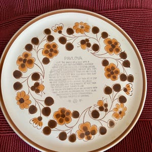 May include: A round ceramic plate with a cream-coloured base and a brown rim. The border features a floral pattern in shades of orange and brown. The centre contains a printed recipe for Pavlova, including instructions for ingredients and baking.
