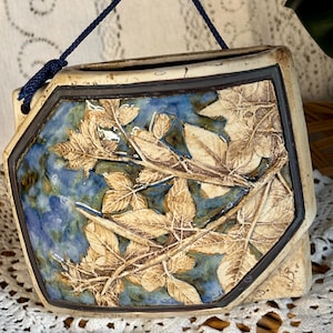 May include: A ceramic wall pocket planter featuring a raised relief of beige leaves against a mottled blue and green background. The piece has a dark brown border and a blue cord attached to the top for hanging, with an open top for plant placement.