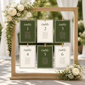 May include: A light wood frame holds six rectangular seating chart cards suspended by wooden clips. The cards alternate between dark green and cream backgrounds, each labeled table 1 through 6 with guest names and white floral accents.