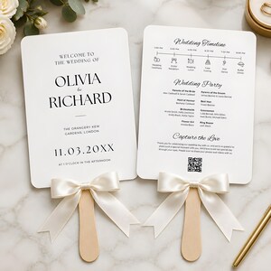 May include: Two white rectangular wedding programme fans with rounded corners, featuring black text and a wooden handle adorned with a cream satin ribbon bow. The front displays names, date, and location, while the back shows a timeline and wedding party list.