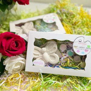 May include: White boxes with butterfly-shaped paint-your-own kits. Each kit includes a ceramic butterfly, paint pots, and a paintbrush. The boxes are displayed on a bed of colorful shredded paper and near red and white roses. The text on the box reads "Paint - Create - Enjoy".