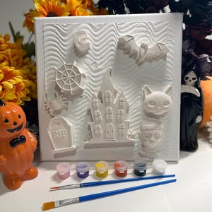 May include: A square white canvas featuring raised relief shapes of a haunted house, a bat, a spider web, a tombstone with the text RIP, a cat, and a monster head. Below the canvas are six small paint pots in various colors and two paintbrushes.
