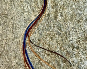 May include: A collection of colorful hair feathers, including blue, red, and tan, arranged in a flowing, curved pattern. The feathers are displayed against a textured, gray stone surface, creating a natural and artistic look.