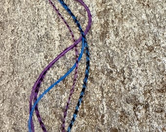 May include: A collection of colorful hair feathers in shades of blue, purple, and black. The feathers are arranged in a flowing, wavy pattern against a textured, light brown surface. These hair accessories add a touch of color and flair.