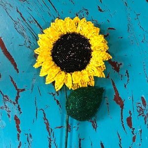 Puede incluir: Un girasol de cuentas con un centro amarillo y hojas verdes. El girasol está sobre un fondo azul.
