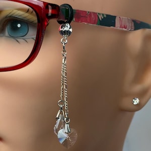 May include: A close-up of eyeglasses with a decorative chain. The red-framed glasses have floral patterned arms. A silver chain with two clear crystal charms, one teardrop and one heart-shaped, hangs from the frame.