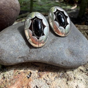 May include: A pair of oval-shaped silver earrings with a central black stone. The earrings have a scalloped edge design around the black stone, creating a unique and elegant look. These earrings are perfect for adding a touch of sophistication to any outfit.