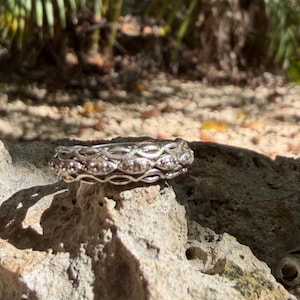 May include: A silver-toned ring featuring a multi-layered band design with an intricate, repeating openwork pattern. The metal surface has a polished finish, and the structure consists of three interconnected rows that create a textured, woven appearance.