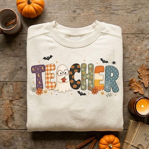 May include: A cream-colored crewneck sweatshirt featuring the word TEACHER in large, patterned letters. The letter A is replaced by a ghost wearing glasses and holding an apple. The design includes bats, pumpkins, flowers, and spiderwebs.