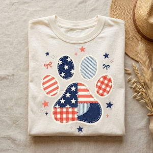 Op de afbeelding: Crèmekleurig T-shirt met een patriottisch pootafdrukontwerp. De pootafdruk bestaat uit verschillende patronen, waaronder sterren, strepen en ruitjes, in rood, wit en blauw. Kleine sterren en strikken omringen de pootafdruk.