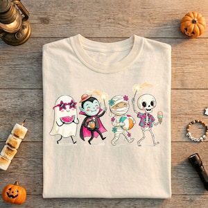 Op de afbeelding: Crèmekleurig T-shirt met een Halloween-thema. Het ontwerp toont stripfiguren verkleed als een spook, vampier, mummie en skelet, elk met feestelijke items zoals watermeloen, snoep, een strandbal en ijs. De karakters lopen in een rij.