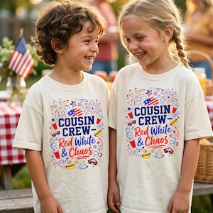 May include: Two children wearing cream-colored t-shirts with a colorful graphic. The graphic includes the text "COUSIN CREW - Red White & Chaos" with fireworks, an American flag, hot dogs, and sunglasses.
