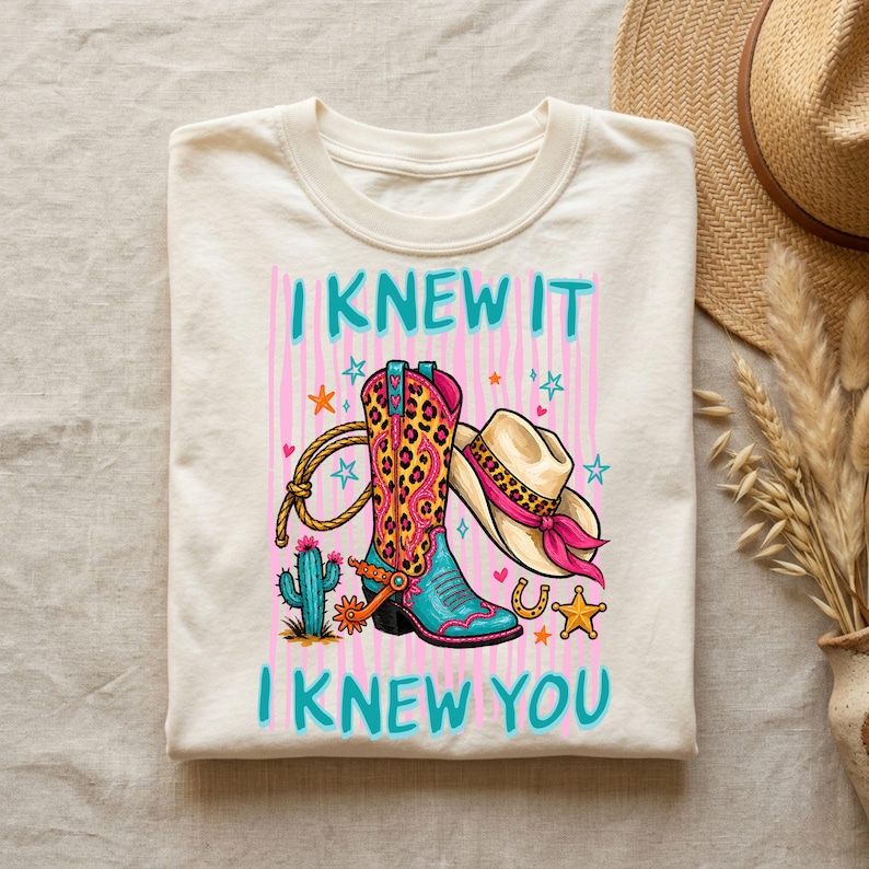 Puede incluir: Una camiseta de color crema con un estampado gr&aacute;fico de una bota de vaquero con estampado de leopardo, un sombrero de vaquero con una banda rosa, un lazo, un cactus y estrellas. El texto "I KNEW IT" y "I KNEW YOU" est&aacute; impreso en azul cerceta.