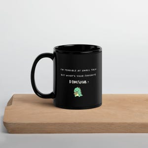 May include: A black ceramic mug featuring the white text "I'M TERRIBLE AT SMALL TALK BUT WHAT'S YOUR FAVORITE DINOSAUR?" and a small, green, cartoon-style dinosaur illustration holding a brown cookie centered on the front surface.