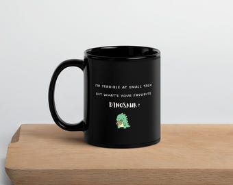 May include: A black ceramic mug featuring the white text "I'M TERRIBLE AT SMALL TALK BUT WHAT'S YOUR FAVORITE DINOSAUR?" and a small, green, cartoon-style dinosaur illustration holding a brown cookie centered on the front surface.
