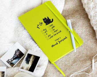 May include: A lime green mood journal with a black cat illustration and the text "F*ck" and "Mood Journal". The journal has a matching elastic band and bookmark. A light blue pen and several polaroid photos are next to the journal.