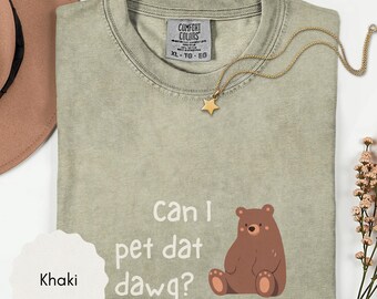 May include: A khaki-colored crew neck t-shirt featuring a graphic of a brown bear and the white text that reads, Can I pet dat dawg? The shirt includes a Comfort Colors brand label at the inner neckline and is paired with a gold star pendant necklace.