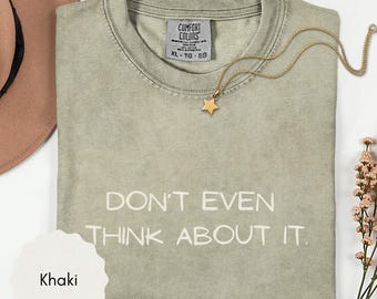 May include: A khaki-colored crew neck t-shirt with the text "DON'T EVEN THINK ABOUT IT." printed in white. The shirt features a Comfort Colors brand label at the inner neckline and is displayed with a gold star pendant necklace.